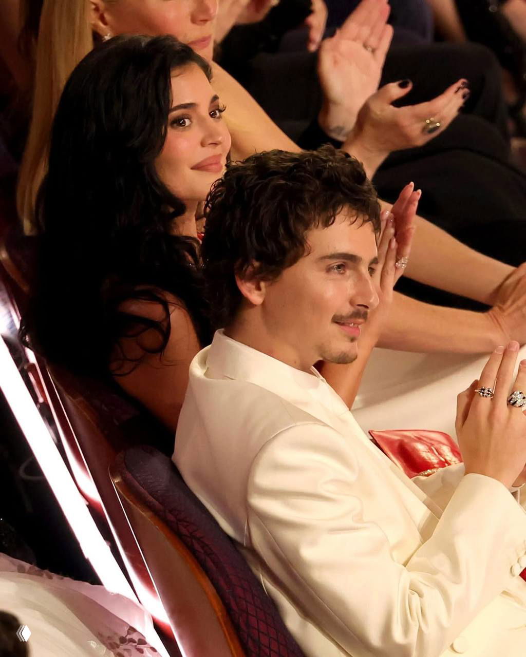 Мужчина в белом костюме аплодирует, рядом женщина с тёмными волосами наблюдает; кадр снят в зале во время награждения.