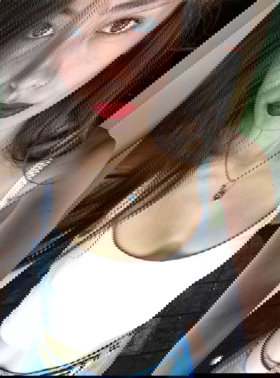 Селфи молодой женщины в белой майке: видна верхняя часть тела, ожерелье с оберегом и яркая помада — динамичный кадр.