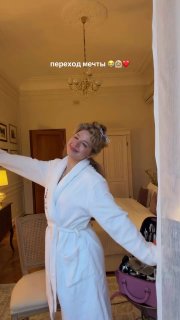 Девушка в белом халате улыбается и распахивает руки в светлом гостиничном номере; на заднем плане видны кровать, кресло, люстра и сумка.