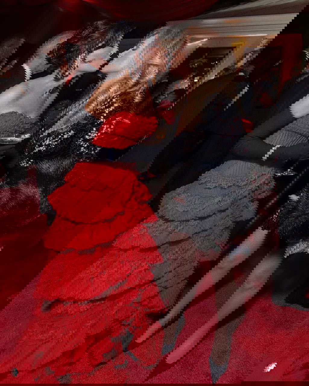 Крис Дженнер целуется в щёку с гостьей в праздничной обстановке: красный ковер, гости в вечерних нарядах и бокалы с шампанским в руке.