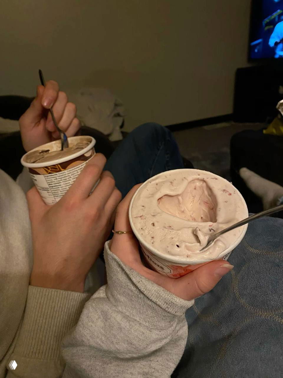 Пара ест мороженое из стаканчиков на домашнем фоне; в одном стаканчике видно сердечко из мороженого — уютный совместный кадр.