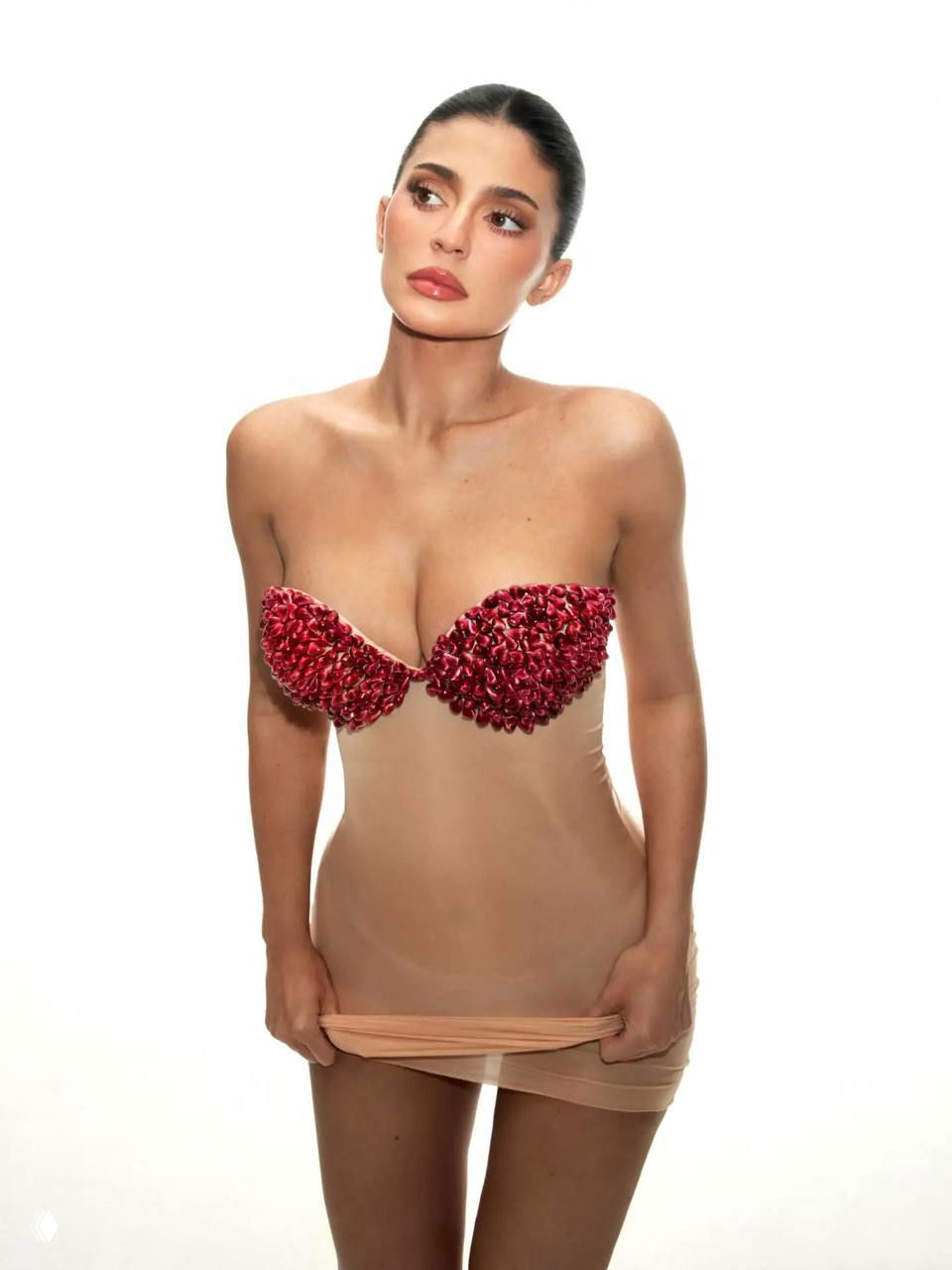 Полнотелая фотография: Кайли в полупрозрачном телесном платье с декором из семян граната на груди, нейтральный фон, взгляд в сторону.