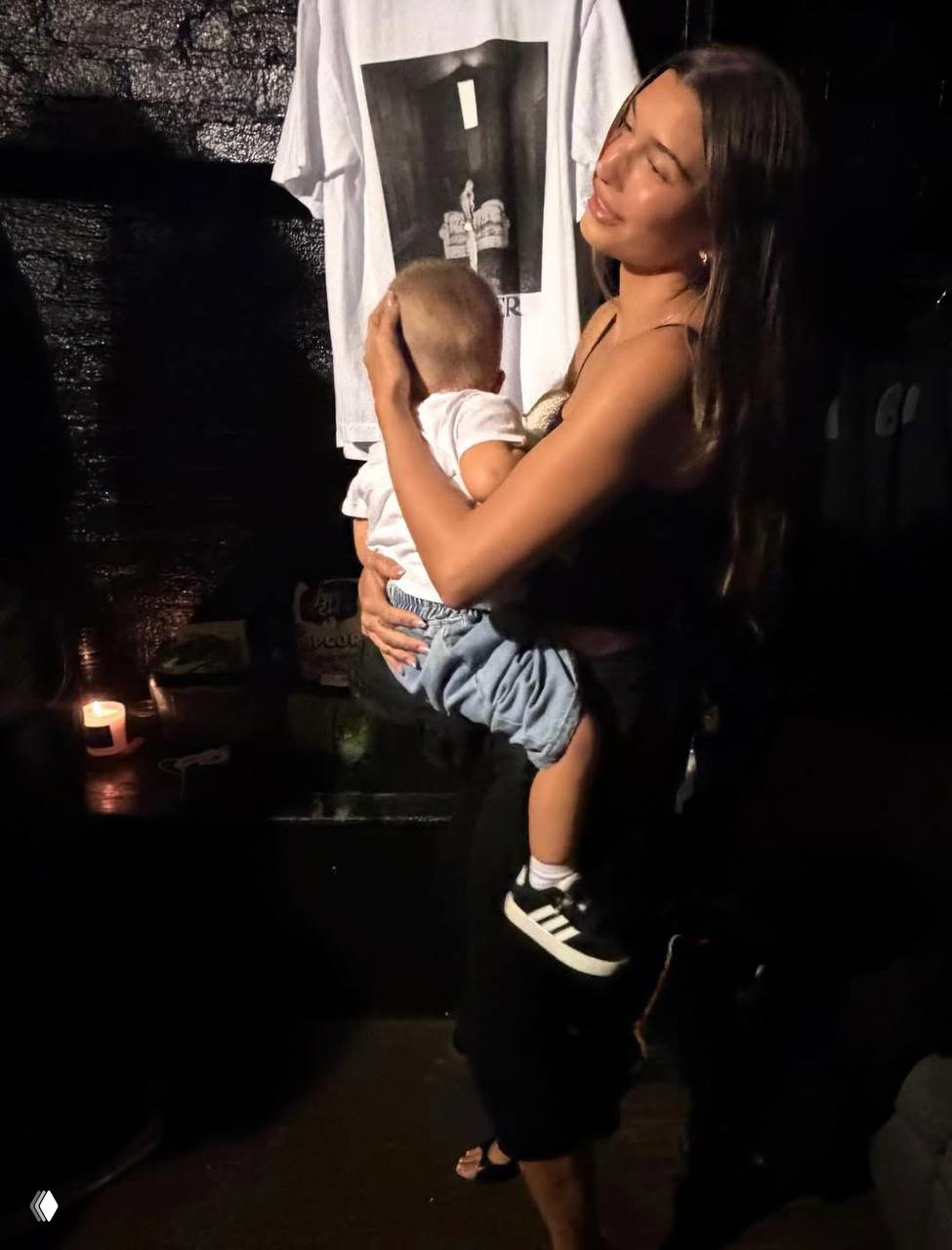 Женщина в тёмном наряде обнимает маленького ребёнка на руках в полумраке — тёплая семейная сцена на тёмном фоне.