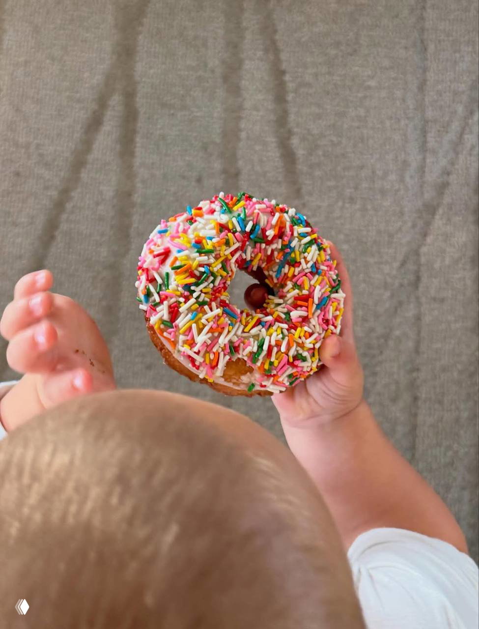 Младенец держит пончик с цветной посыпкой в руках, вид сверху на голову ребёнка и лакомство.