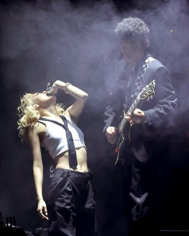 Исполнительница в эмоциональном моменте выступления наклоняется к микрофону рядом с гитаристом в дыму сценического оформления.