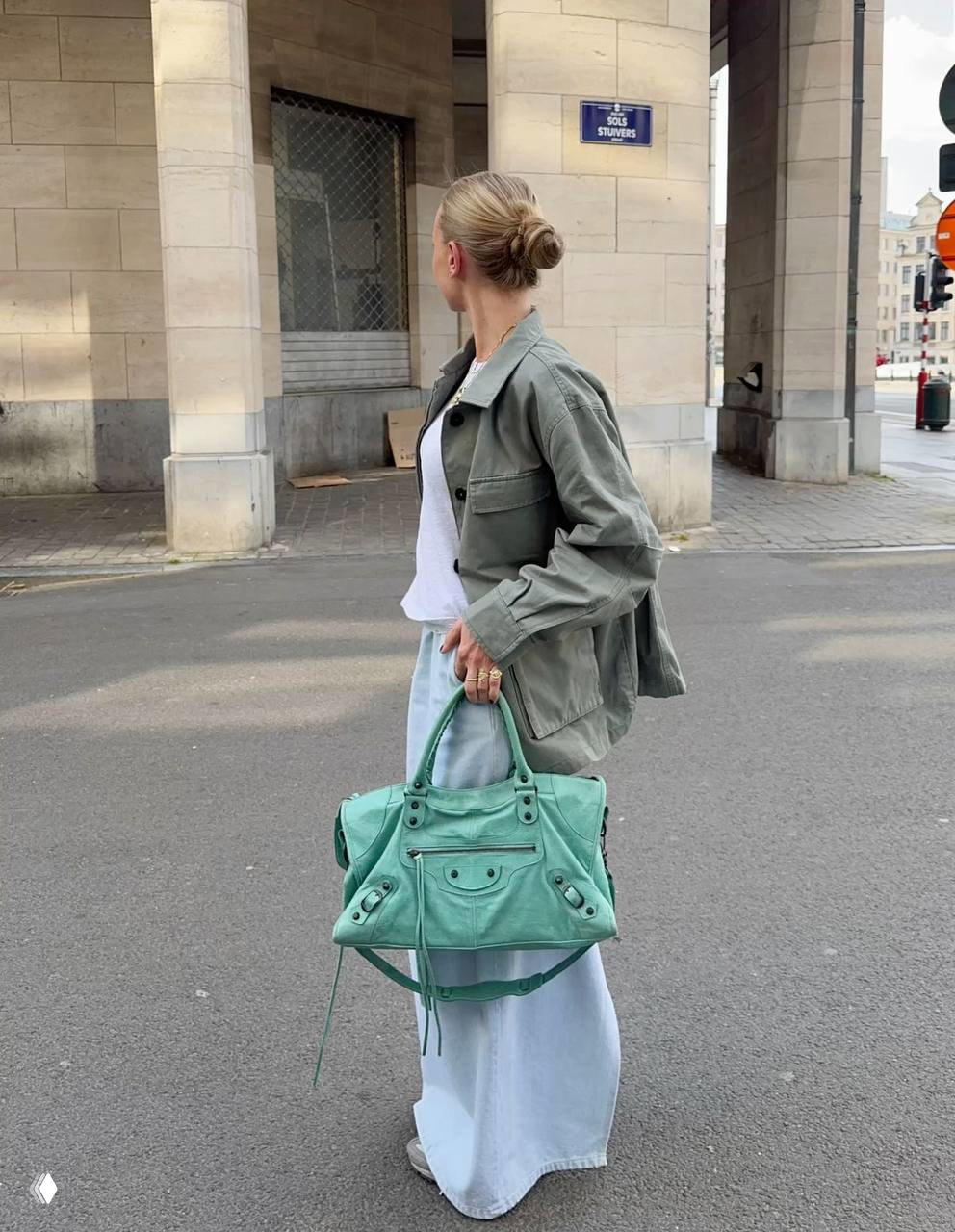 Уличный образ: женщина в мятной сумке и оливковой куртке, длинная джинсовая юбка и повседневная композиция аксессуаров на фоне городской архитектуры.