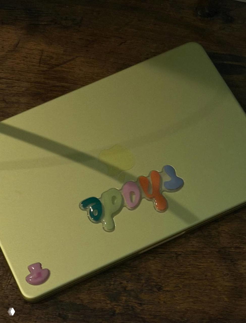 Зелёная крышка ноутбука с объёмными разноцветными наклейками на деревянном столе при мягком освещении.