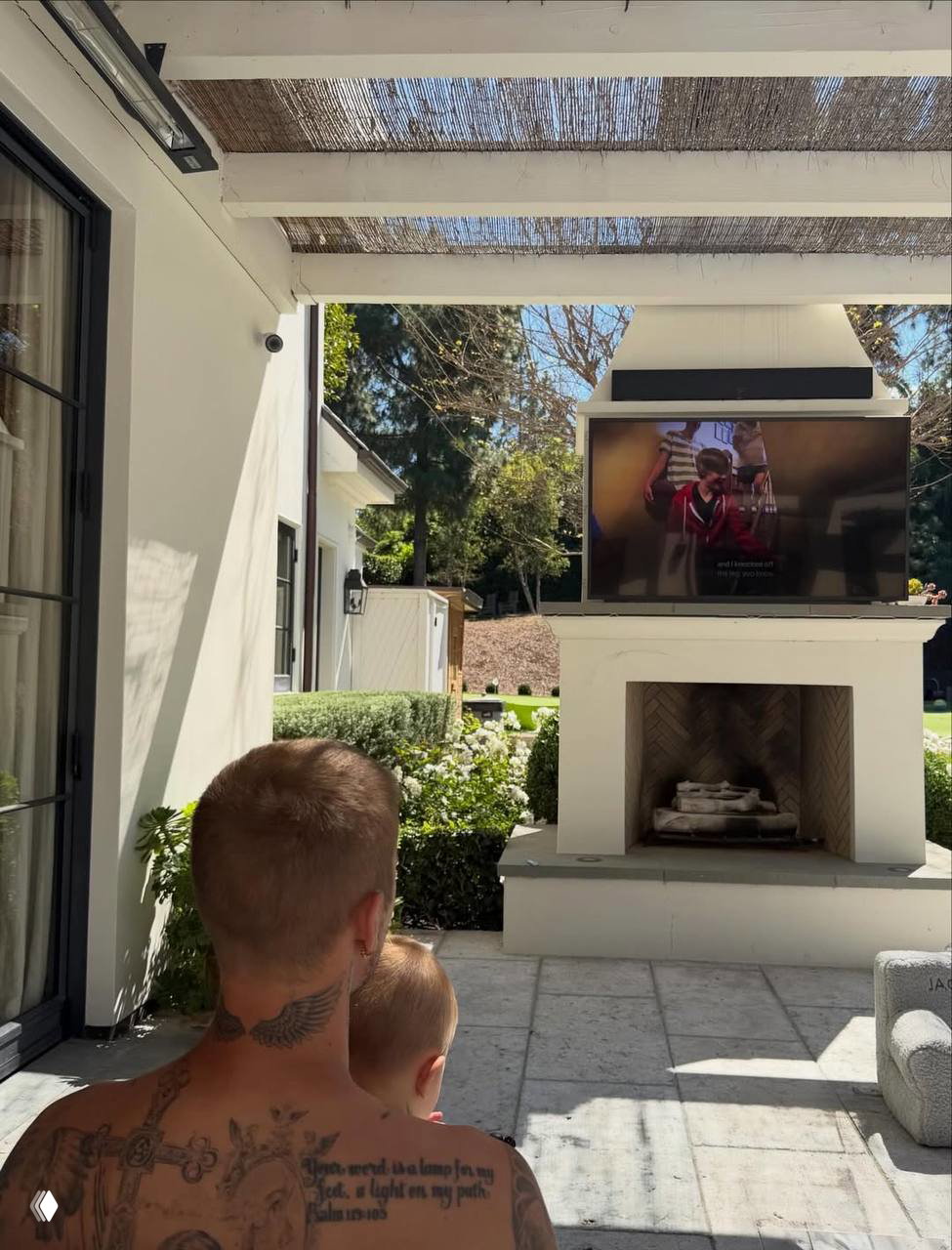 Уличная зона с камином: мужчина со спины держит ребёнка на руках и смотрит телевизор на открытом воздухе в солнечный день.