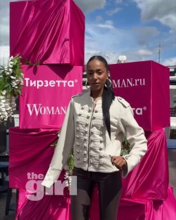 Алиша в светлом гусарском жакете с крупными пуговицами на фоне ярко‑розовых декораций и логотипа Woman.ru во время съемки.