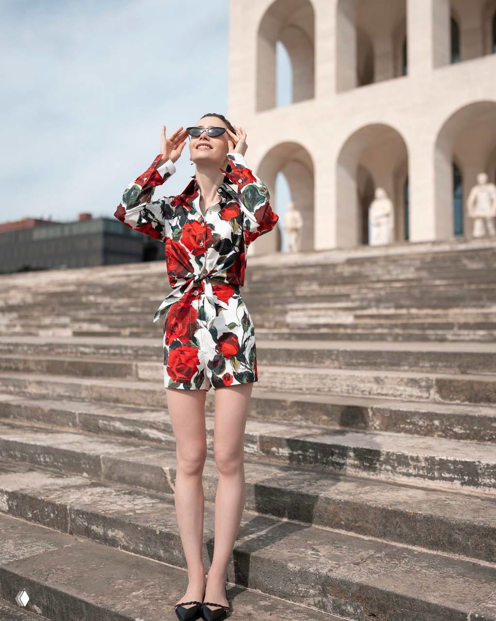 Героиня в коротком костюме с крупными красными цветами стоит на широких каменных ступенях рядом с архитектурным памятником, солнце и тени.