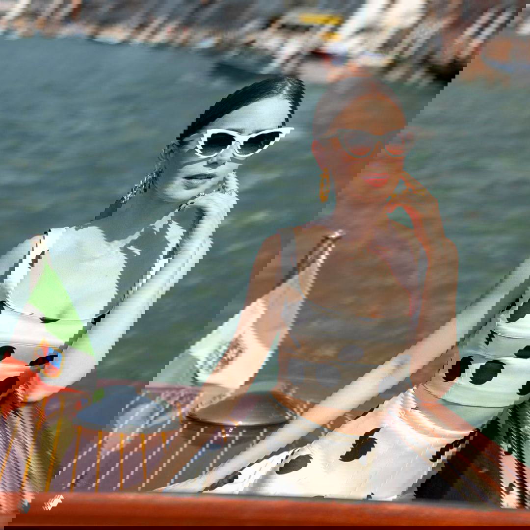 Героиня в белом топе и юбке с черным горошком позирует на борту лодки у воды; виден флаг страны и морской пейзаж на заднем плане.