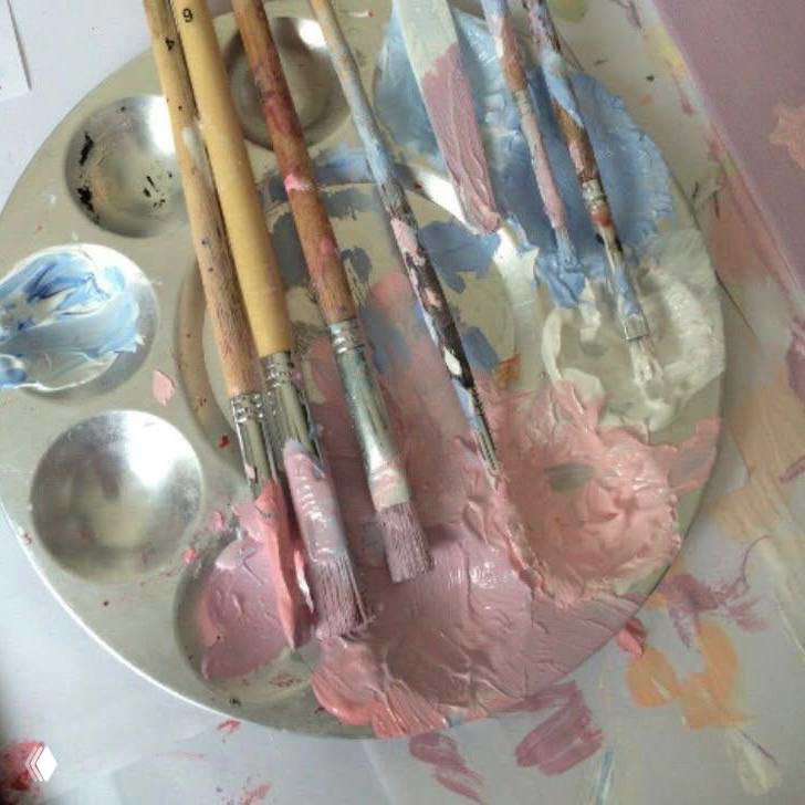 Палитра с розовыми красками и кистями на белой поверхности; мазки и текстура краски создают творческую студийную атмосферу эстетического референса.