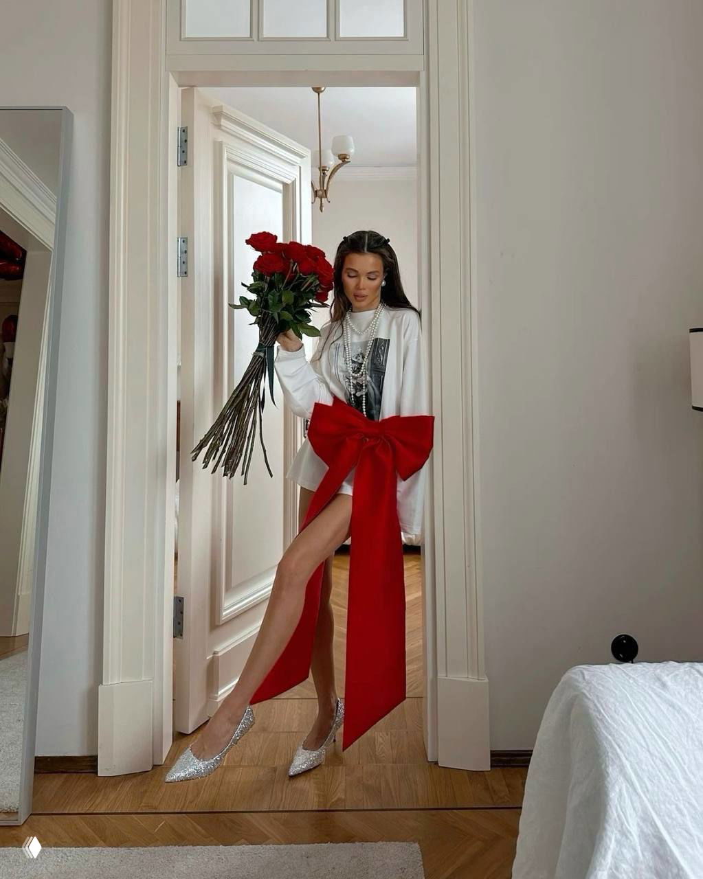 Модель в длинной белой рубашке с большим красным бантом и букетом роз — праздничный, театральный образ с ярким красным акцентом и серебристой обувью.