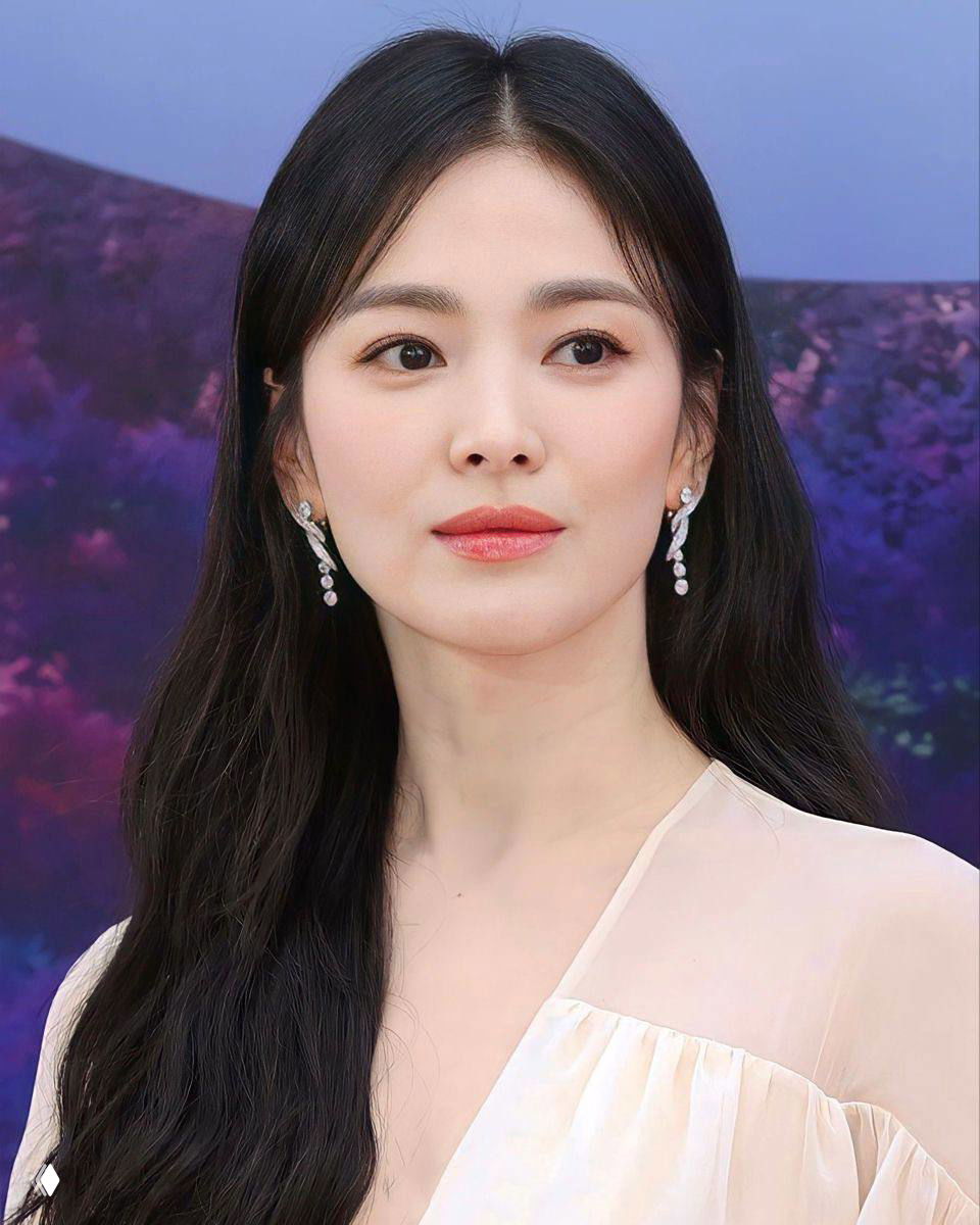 Портрет Сон Хе Гё на мероприятии: длинные тёмные волосы, гладкая сияющая кожа и аккуратные серьги на фоне тёмно-фиолетовой сцены.