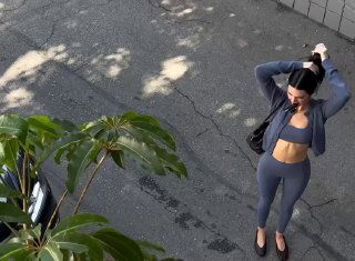 Маленький превью‑кадр: вид сверху на женщину в комплекте для тренировок, собирающую волосы в пучок, фон — асфальт и листья растений у бордюра.