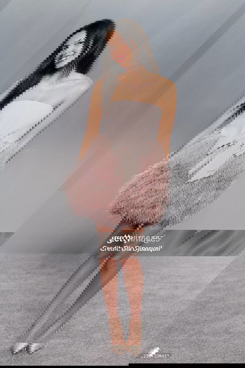 Короткое светло‑розовое платье с пышной перьевой юбкой и бантом, образ в коктейльном стиле на серой фотозоне мероприятия.