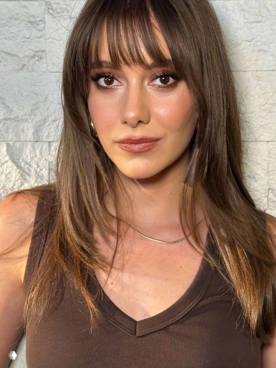 Крупный план молодой женщины с мягким макияжем и чёлкой на светлой мраморной стене; фокус на глазах и естественном сиянии кожи при естественном свете.