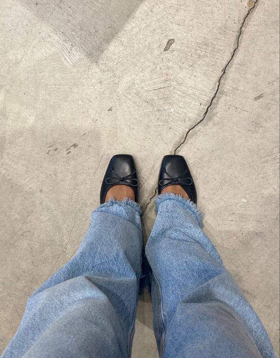 Ноги в чёрных балетках и джинсах стоящие на трещиноватом бетонном полу — минималистичный ракурс обуви для поездки.