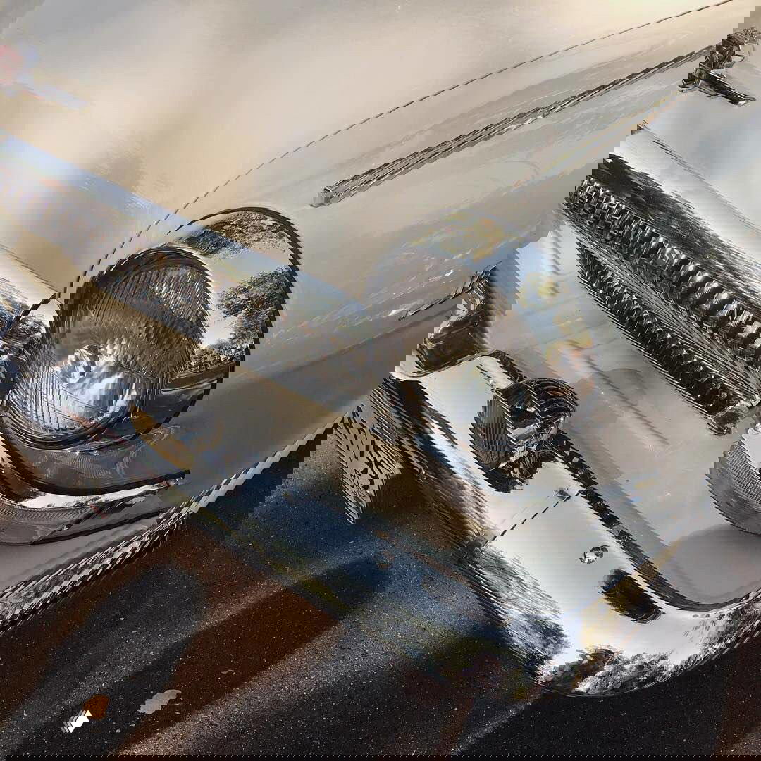 Крупный план хромированной радиаторной решётки и круглой фары классического ретро-автомобиля, отражения улицы на блестящем бампере.