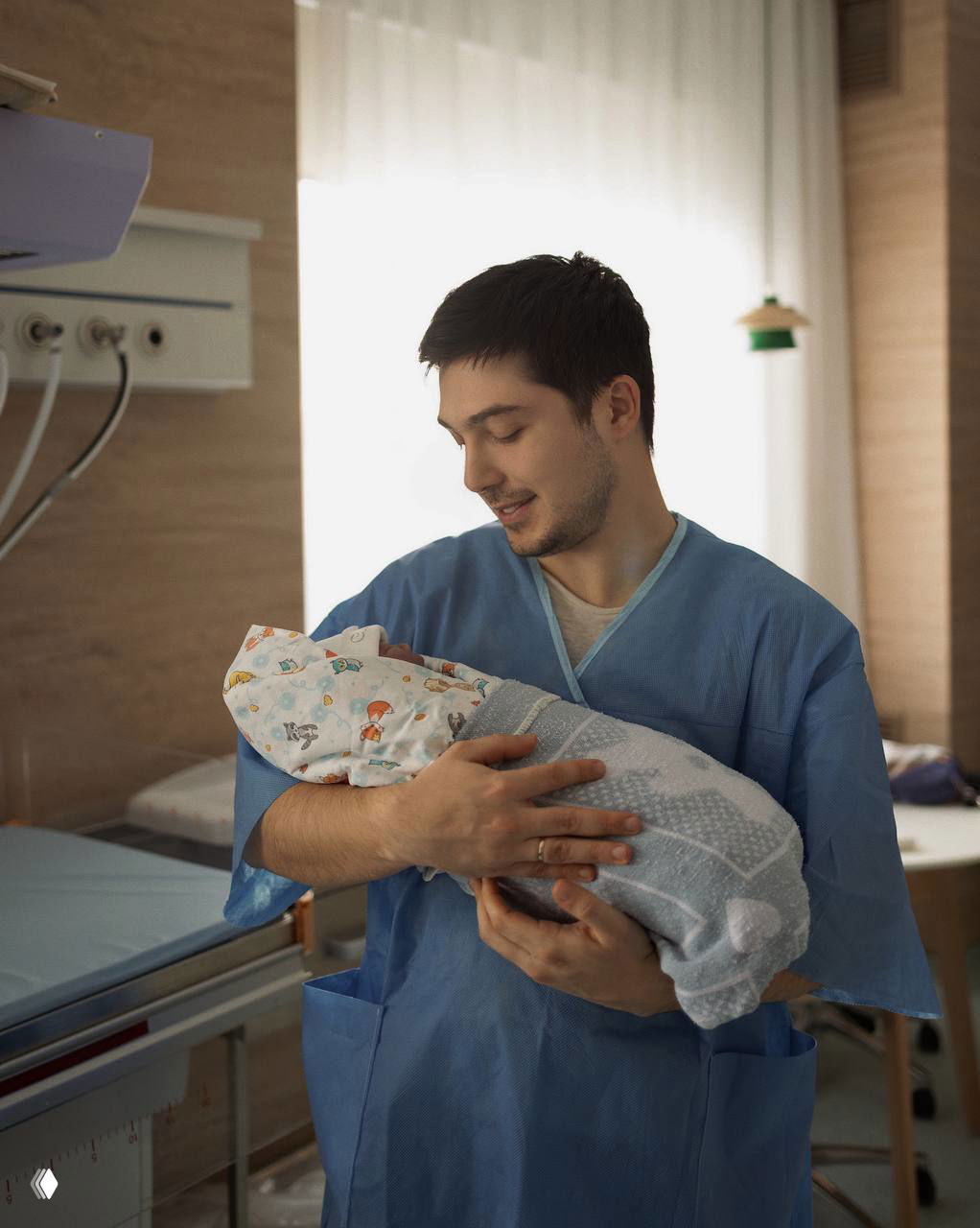 Мужчина в медицинском халате держит завернутого новорождённого в палате роддома; на заднем плане видно оборудование и светлое окно.