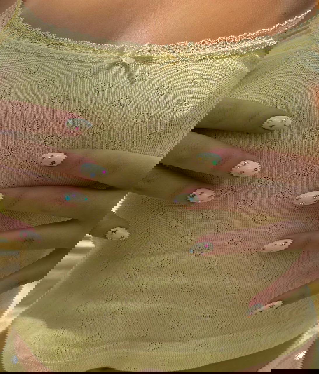 Руки в светло‑жёлтой майке с белым маникюром: на овальных ногтях пришитые разноцветные стразы разных размеров, модный весенний образ.