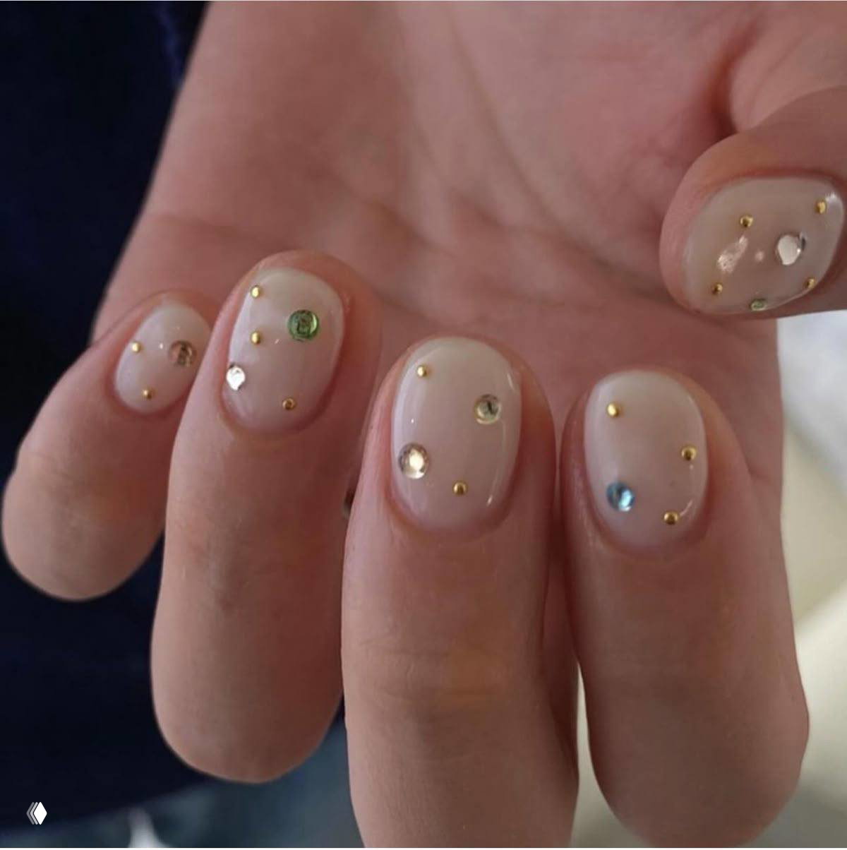 Фотография пальцев с натуральным нюдовым покрытием и цветными стразами: аккуратные круглые камни и золотые акценты на коротких ногтях.