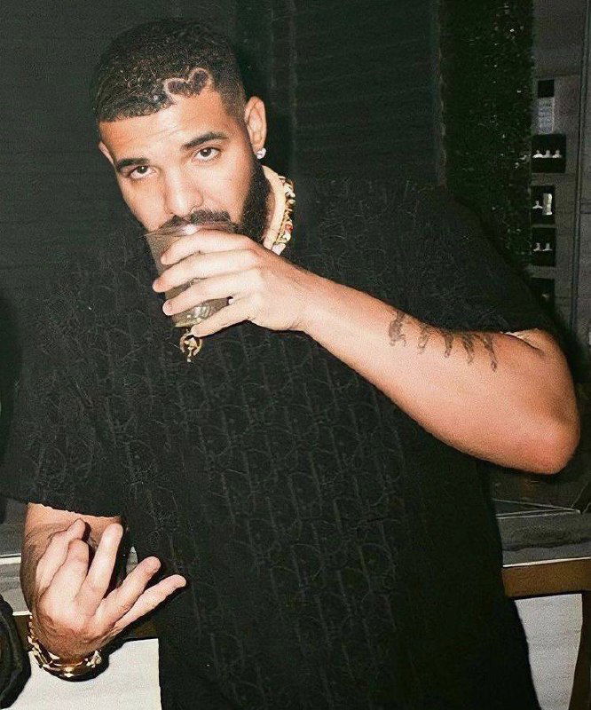 Фотография мужчины с бородой в чёрной футболке, он держит стакан в руке; кадр в интерьере с приглушённым тёплым светом.