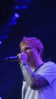 Певец со светлыми волосами на сцене держит микрофон; татуировка на руке, фиолетово‑синее освещение и тёмный фон.