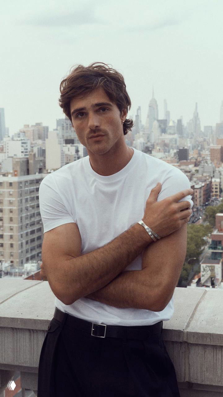 Портрет Джейкоба Элорди в белой футболке, руки скрещены; он стоит на крыше на фоне городского пейзажа с небоскрёбами и серым небом.