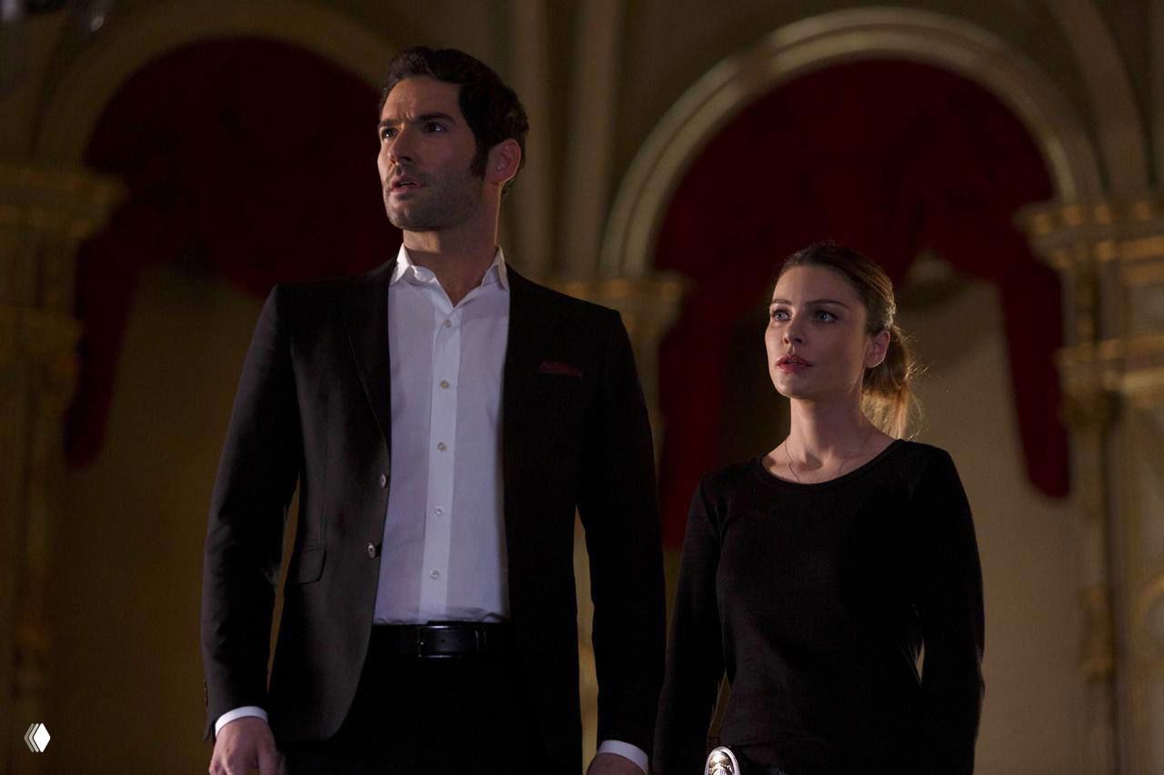 Кадр из сериала «Люцифер»: двое персонажей стоят в театральном интерьере, тёмная одежда и деловой костюм на фоне архитектурных арок и драматичного освещения.