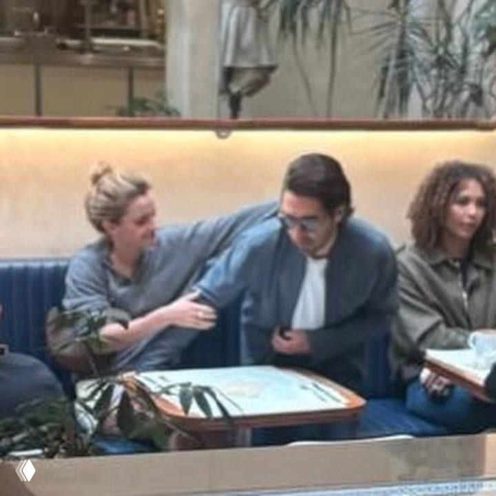 Пара в ресторане в момент рукоприкосновения: Эмма кладёт руку на плечо мужчины, они сидят в кабине рядом с меню и другими посетителями.