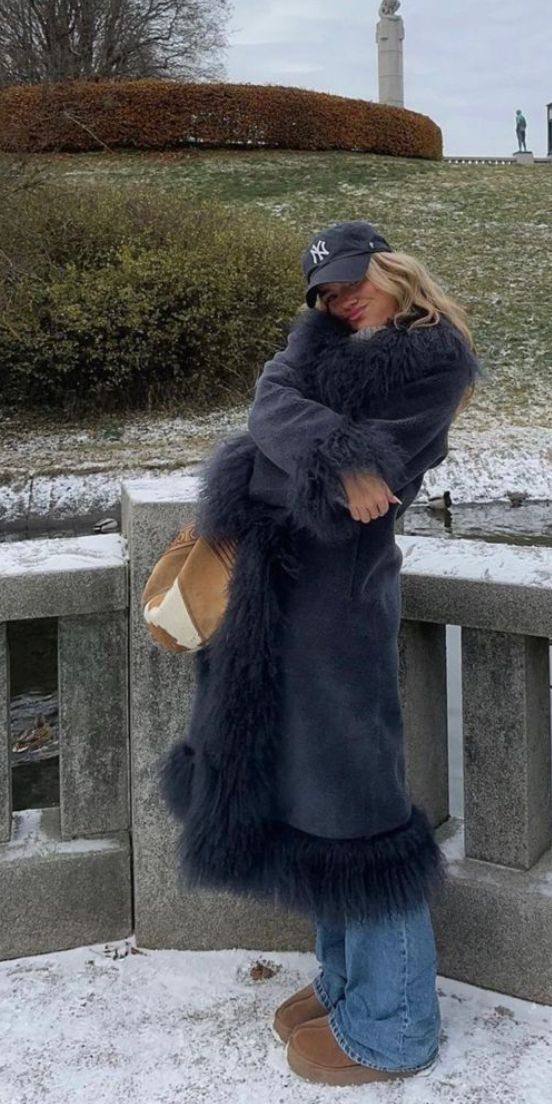 Уличный снимок: женщина в длинном тёмно-синем афганском пальто с пушистыми манжетами на фоне заснеенного городского пейзажа и парковки
