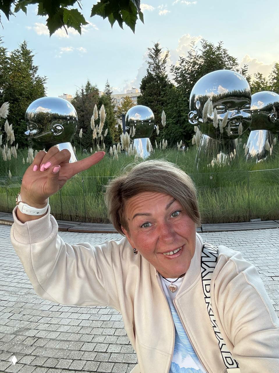 Женщина на фоне парка с зеркальными скульптурами, она улыбается и указывает на арт-объекты в зелёной городской зоне.