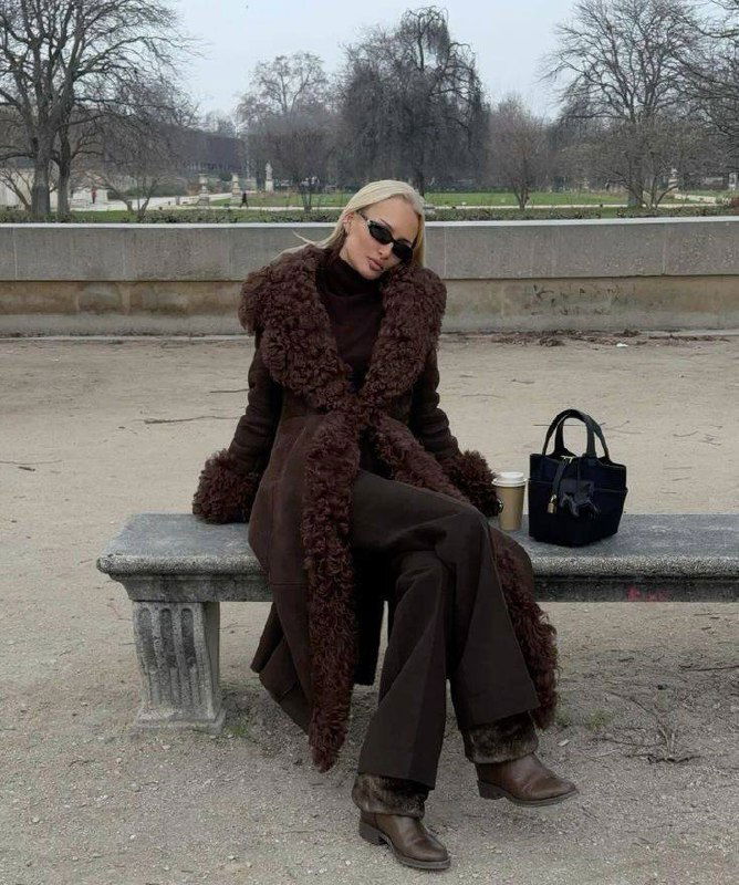 Модель в коричневом длинном пальто с объёмным меховым воротником сидит на скамейке в парке, элегантный осенний образ с солнцезащитными очками