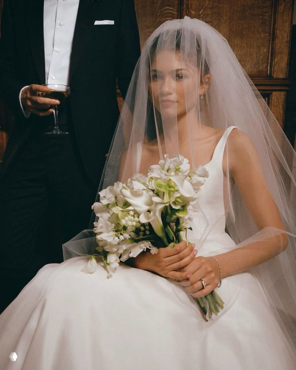 Женщина в свадебном платье с фатой держит букет цветов; рядом частично виден мужчина в смокинге — портретная сцена из фотосъёмки.