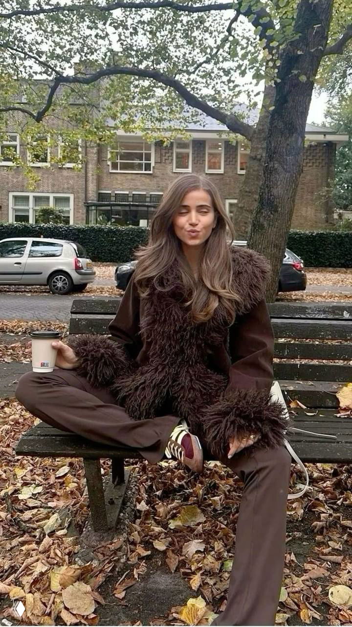 Девушка на скамейке в коричневом пальто с пушистым мехом по краю, рядом сумка и стакан кофе, осенний городской фон с парком