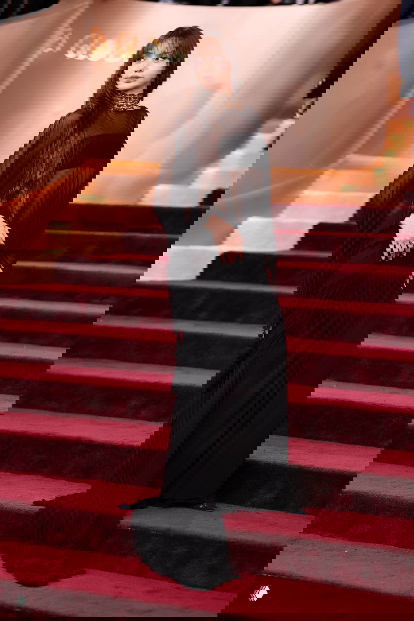 Полный рост: девушка в элегантном чёрном наряде Jacquemus поднимается по ступеням красной дорожки, видна сцена и оформление церемонии.