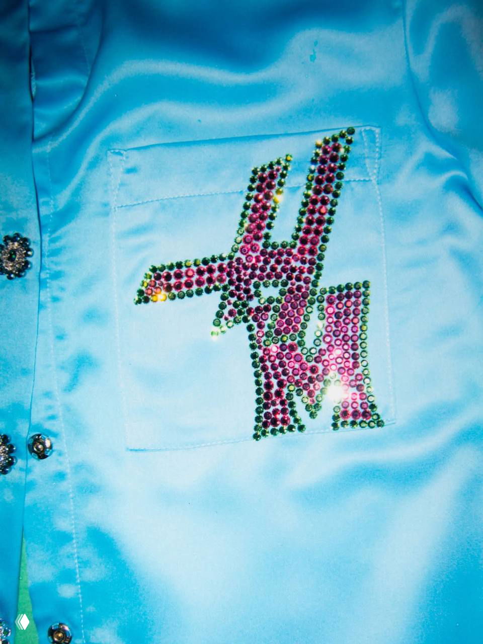 Крупный план голубой сатиновой рубашки с пришитым стразовым логотипом «HM» на кармане, видна деталь сценического костюма.