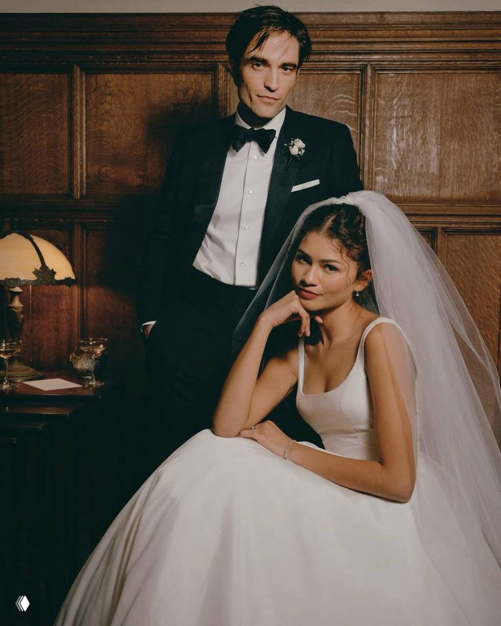 Пара в вечерних нарядах: мужчина в смокинге стоит позади сидящей женщины в свадебном платье — студийная постановочная фотография.