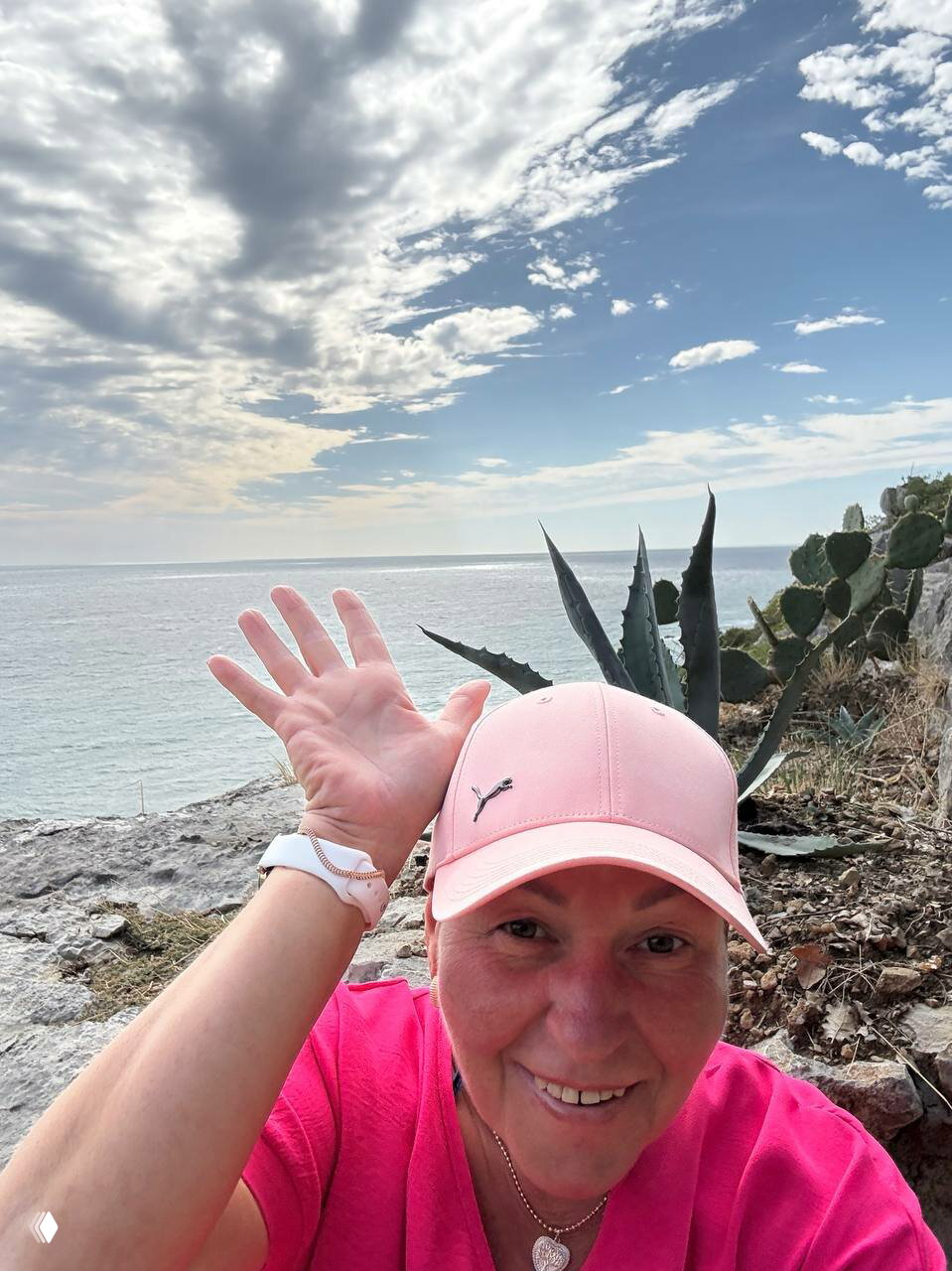 Селфи женщины в спортивной кепке на фоне скалистого побережья и агавы, видно каменистый берег и море за спиной.