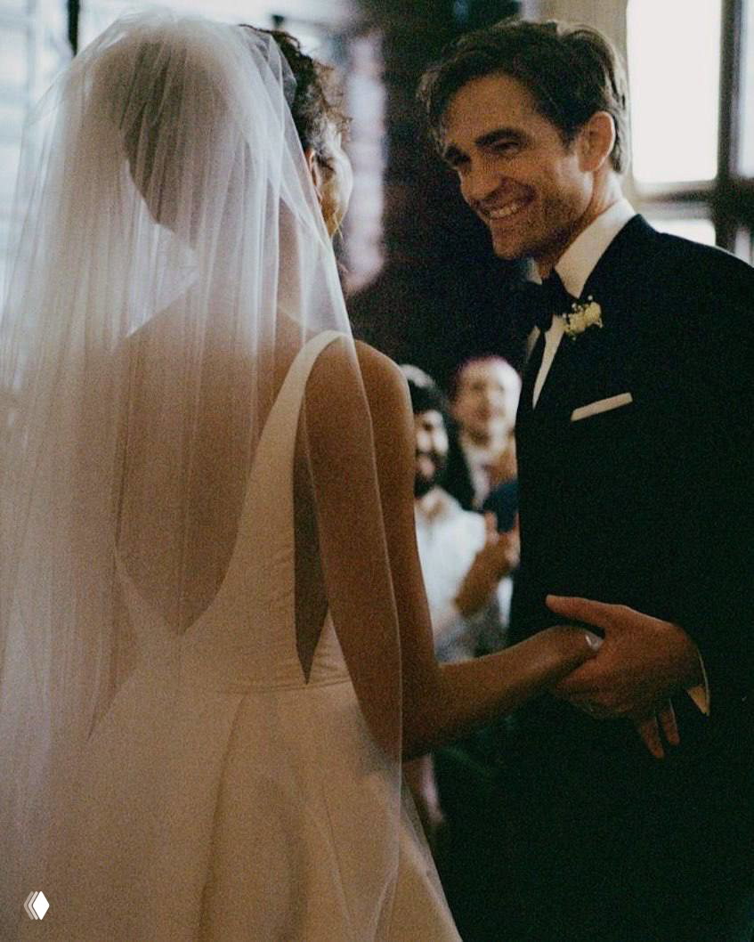 Момент с танцем или поздравлением: женщина в белом платье держит руку мужчины, вокруг гости в полутемном зале — тёплая праздничная сцена.