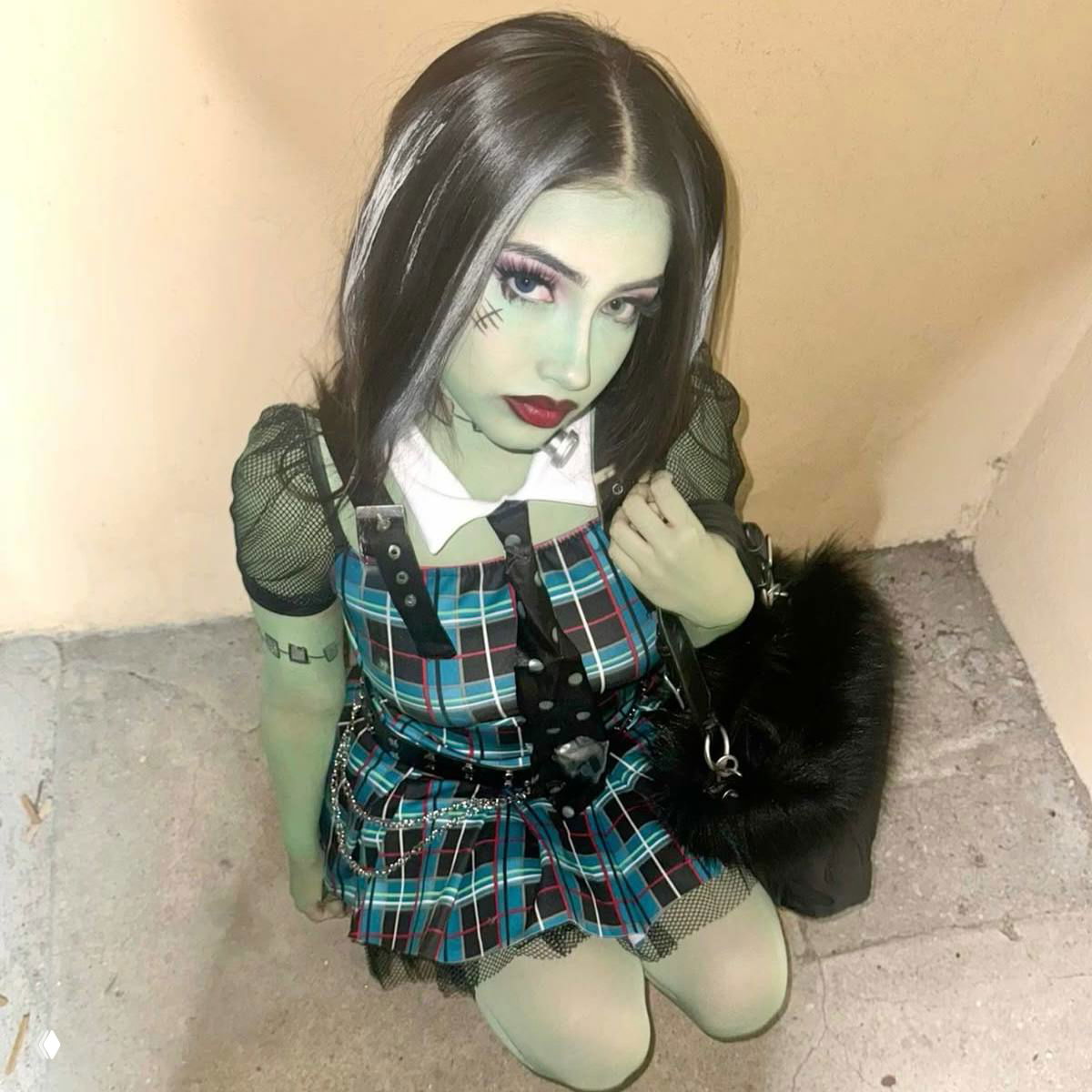Крупный кадр косплеера в образе Фрэнки: зелёный тон кожи, подчёркнутый макияж, чёрные волосы с белыми прядями и клетчатый костюм в стиле школьной формы.