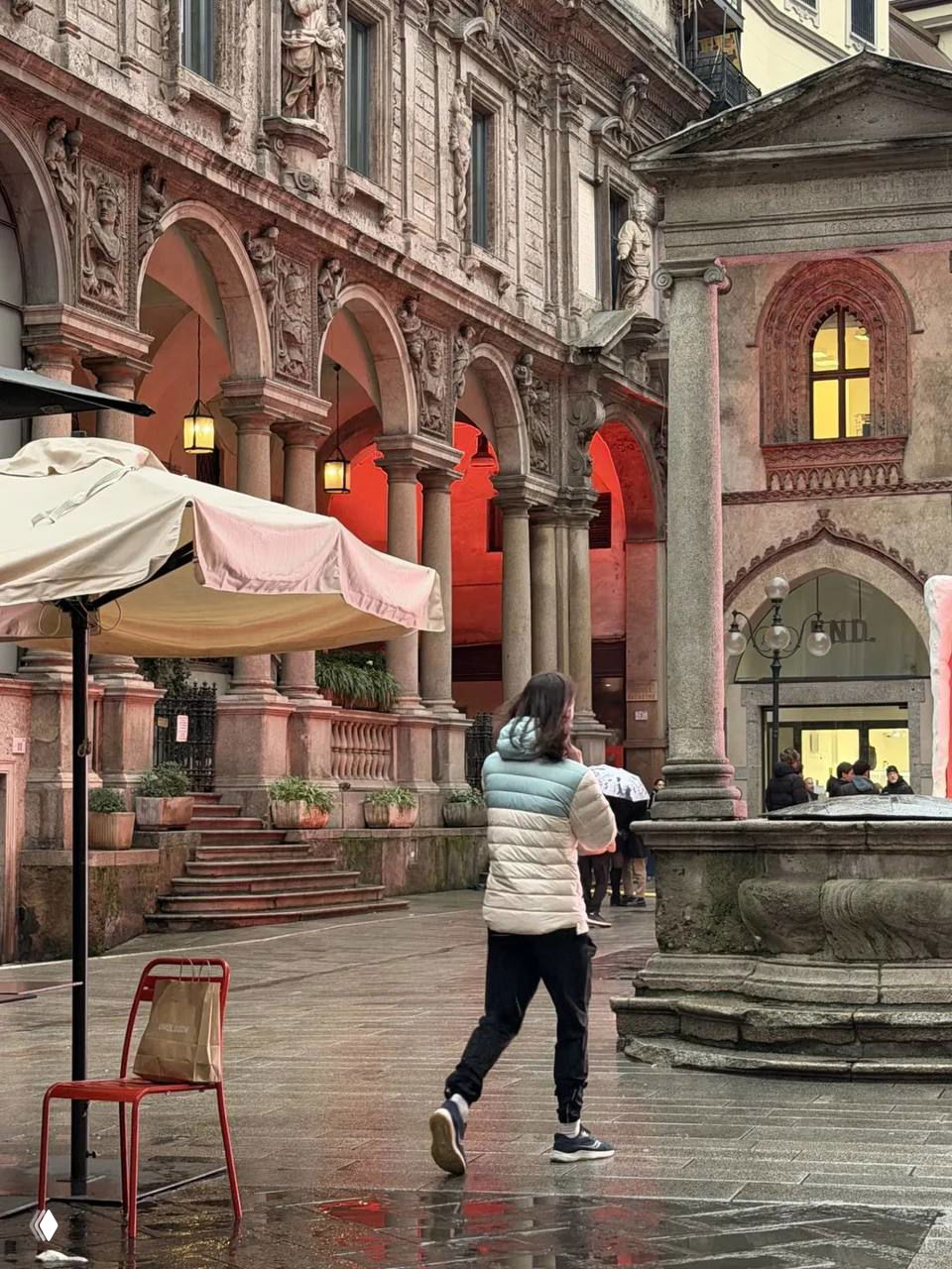Мужчина идёт по историческому дворику в Милане в стёганой куртке, снят сзади; видны арки, фонтан и тёплая подсветка фасадов.