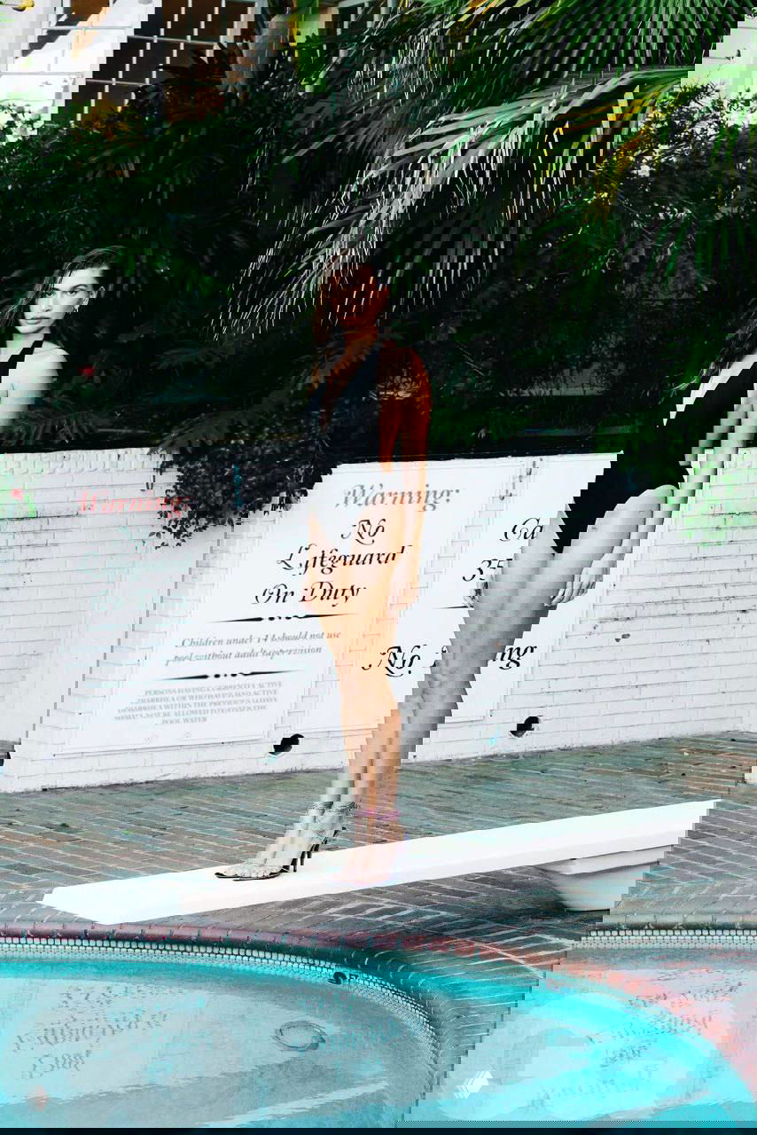 Цветная фотография женщины в чёрном купальнике на трамплине у бассейна; пальмы на фоне, ретро‑модный кадр в стиле фэшн‑съёмки.