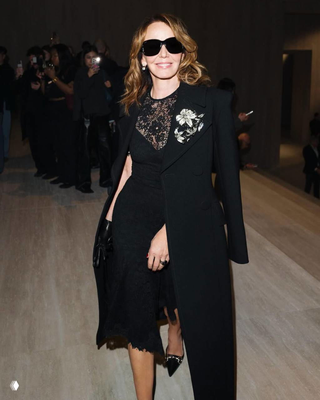 Женщина в черном кружевном платье и пальто выходит на дорожку показа; толпа фотографов на заднем плане фиксирует момент.