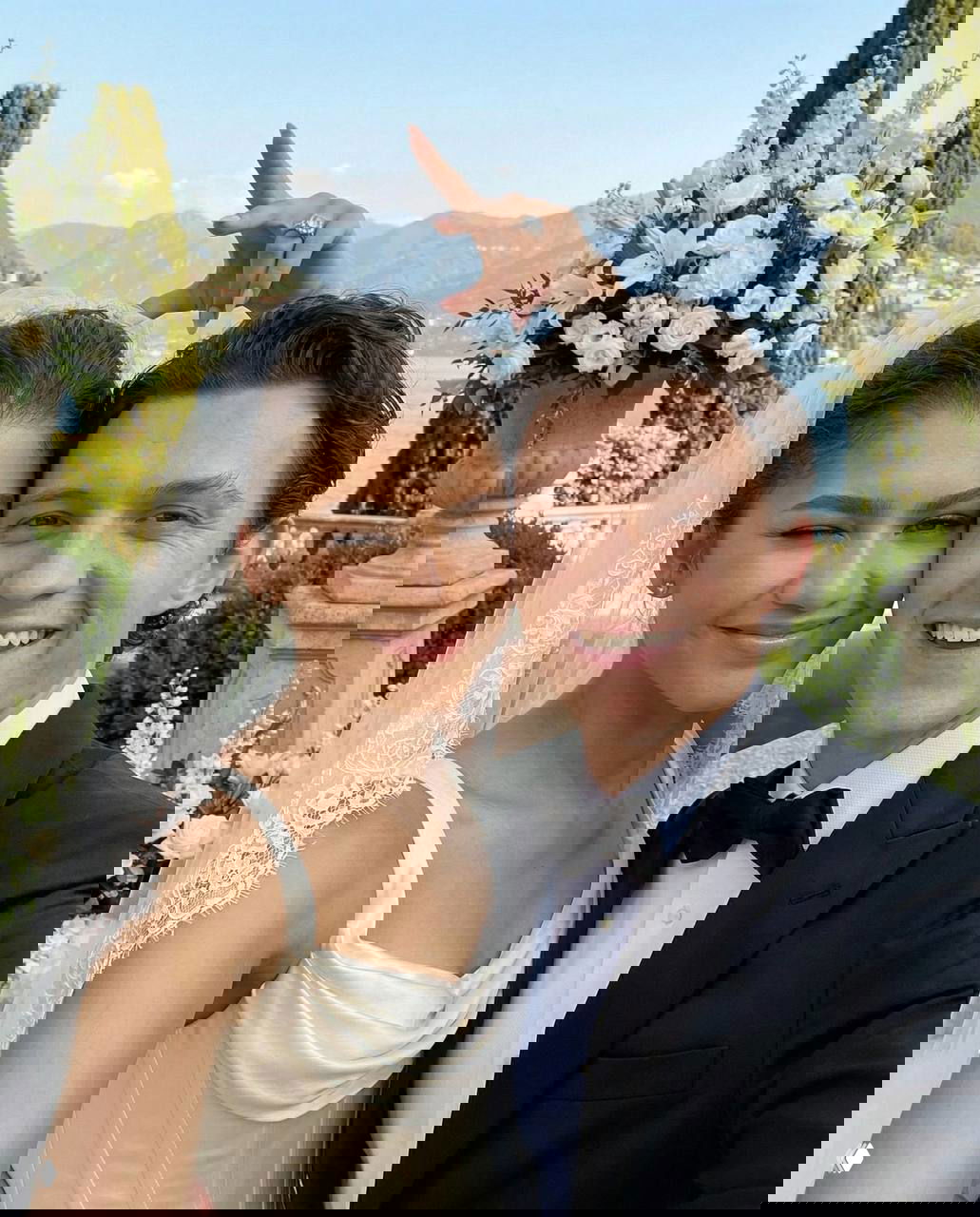 Пара в свадебных нарядах делает селфи на террасе с видом на озеро и горы, вокруг видны цветочные композиции и декор церемонии.