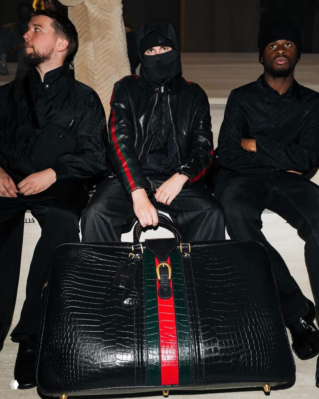 Лица зрителей в первом ряду показа Gucci: трое мужчин сидят рядом, на переднем плане крупная дорожная сумка с фирменной полосой Gucci.