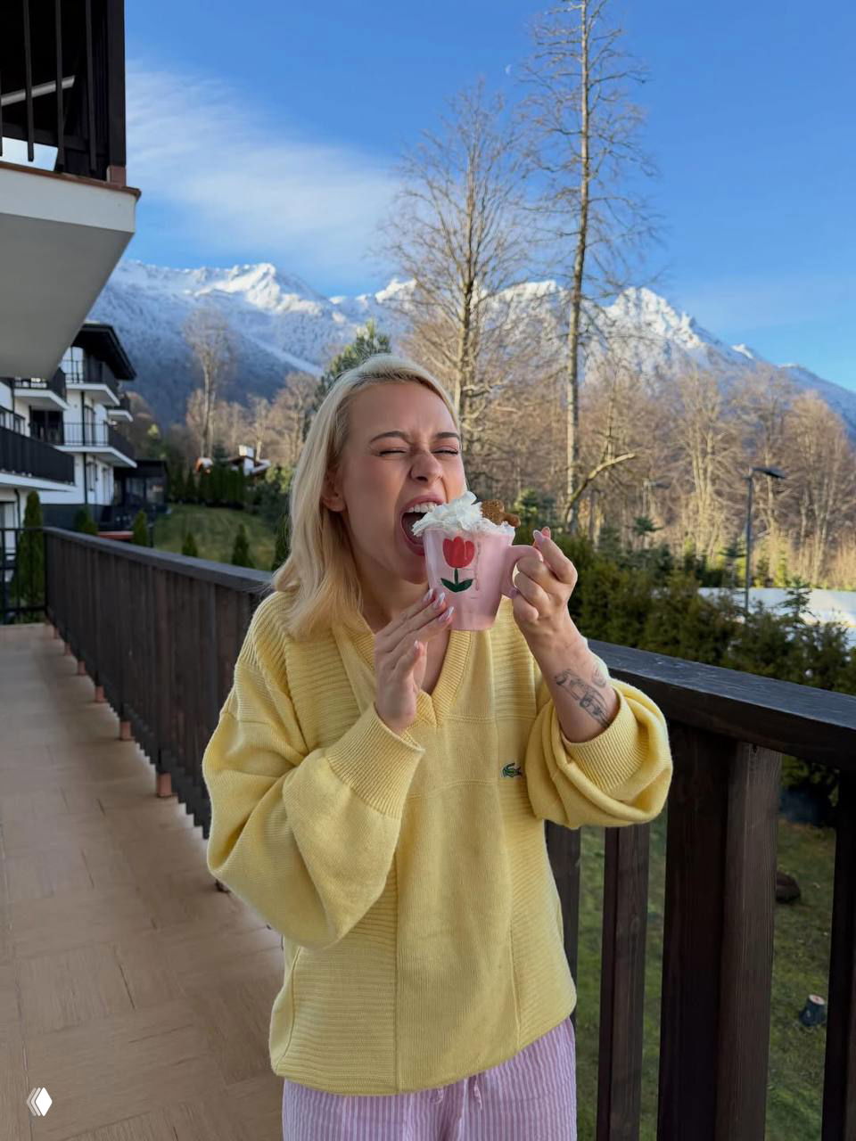 Настя шутливо прикусывает кружку с напитком на балконе курорта; в фоне хвойные деревья и снежные горы, создающие атмосферу отдыха.