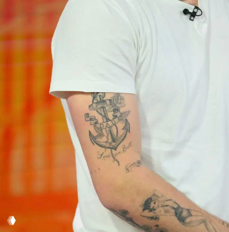 Фрагмент руки с татуировкой якоря и декоративными элементами; снимок показывает тёмные контурные линии, переплетение верёвки и части надписей вокруг рисунка.