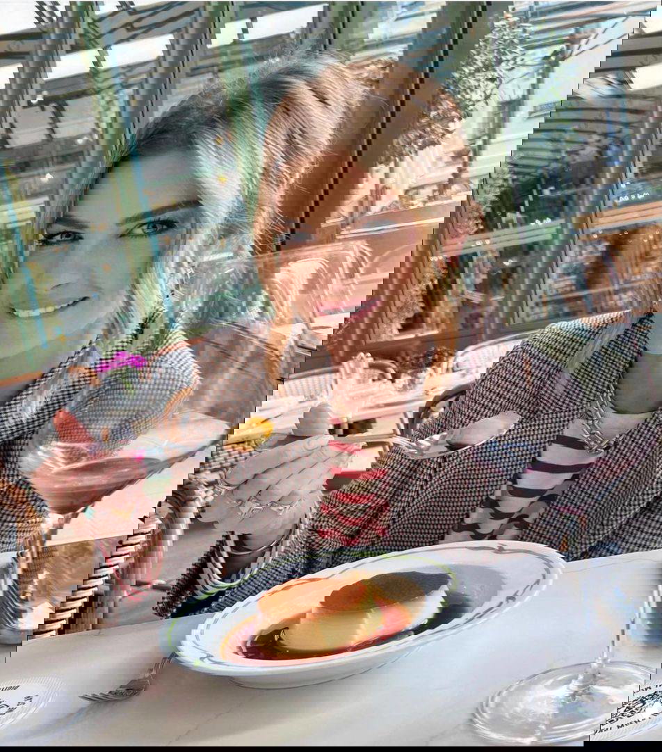 Женщина улыбается за столиком в ресторане, держит ложку над десертом флан; на столе бокал вина и элементы интерьерного декора видны рядом.